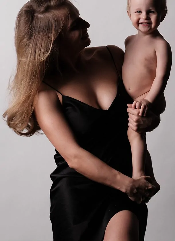 Woman in a black dress holding a smiling toddler, looking at the child with a warm expression against a neutral studio background.