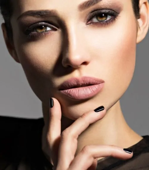 Close-up of a woman with full, well-defined lips and smooth skin, softly contoured makeup, and manicured black nails, with her hand gently touching her chin to highlight lip shape and facial symmetry.